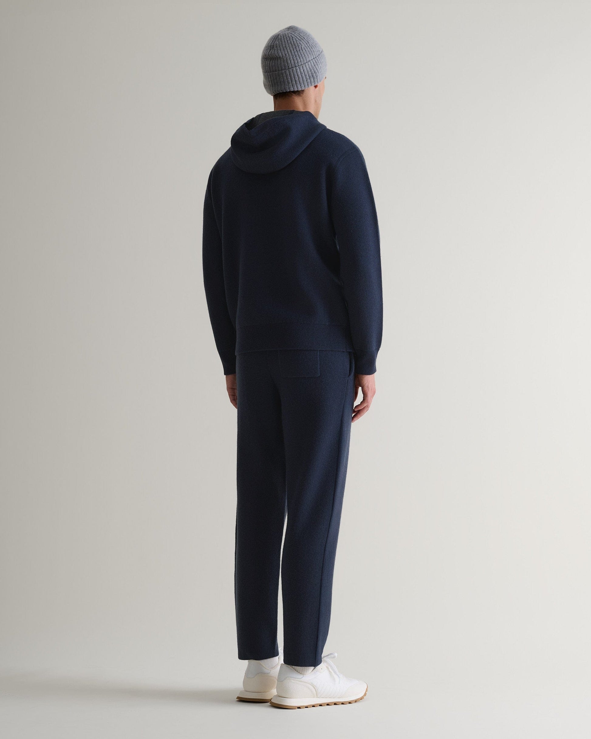 Back view of navy cashmere merino double-faced knitted hoodie with matching trousers, white trainers and grey beanie.
