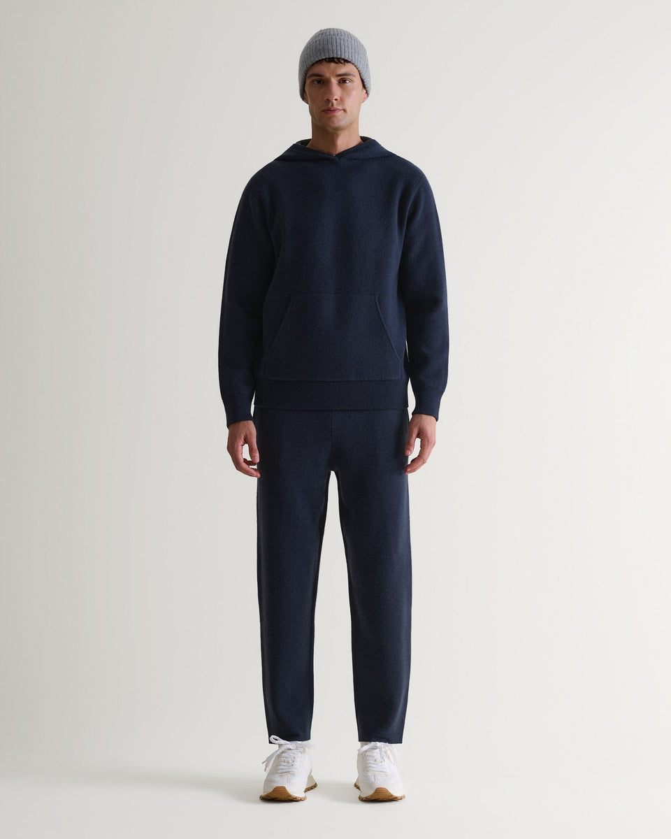 Man wears navy cashmere merino double-faced knitted hoodie with matching trousers, white trainers and light grey beanie.