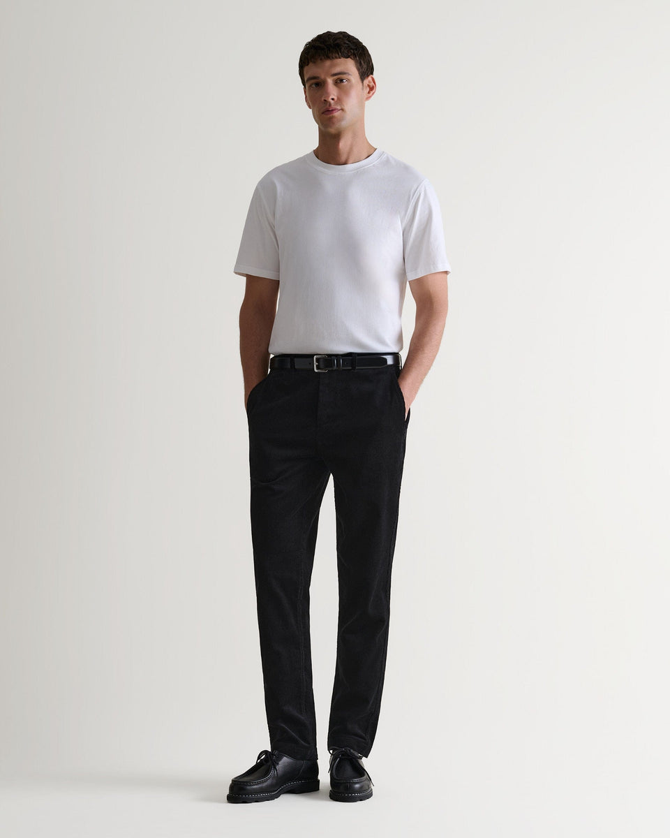 Full length view of man wearing Rise & Fall's black corduroy trousers with white T-shirt and black leather shoes.
