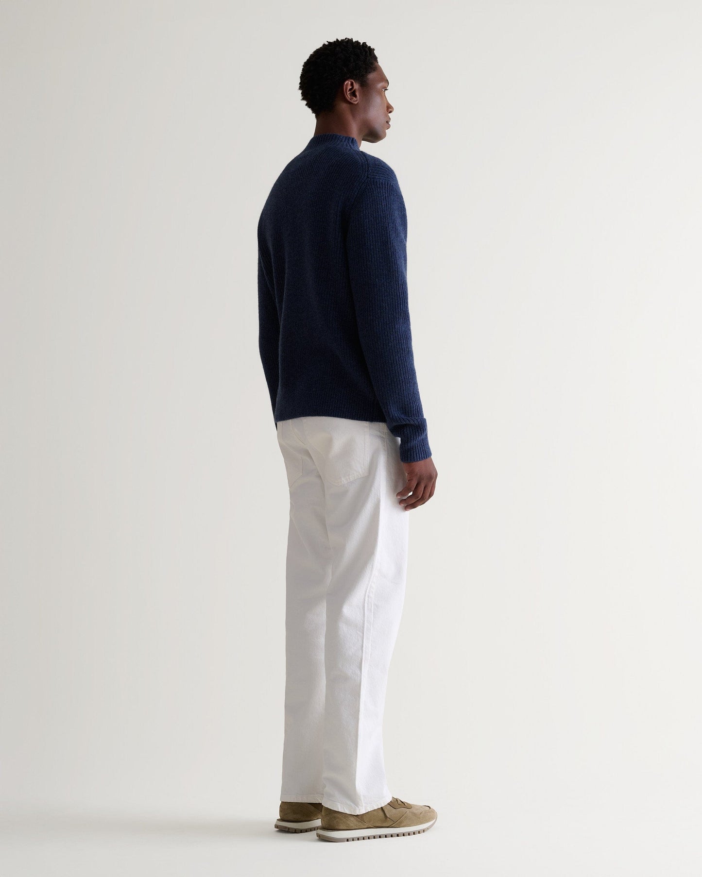 Side view of man wearing Rise & Fall blue cashmere merino stand neck jumper with white trousers and beige suede trainers.