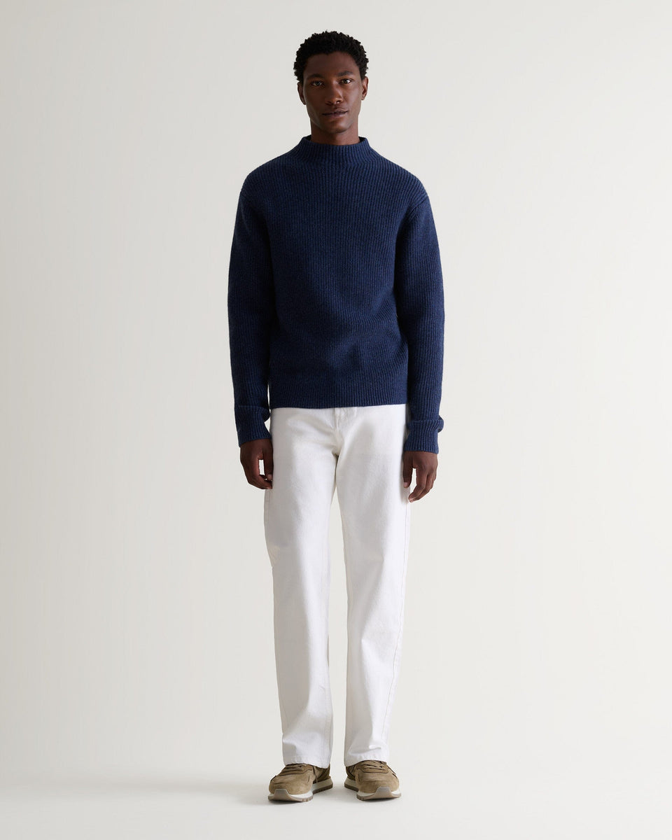 Front view of man wearing Rise & Fall blue cashmere merino stand neck jumper with white trousers and beige suede trainers.