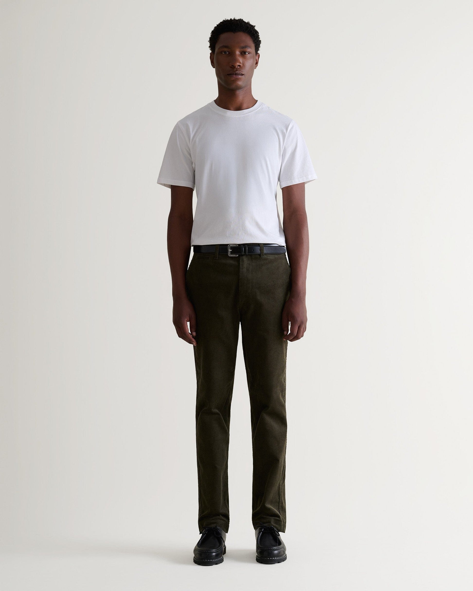Full length front view of man wearing Rise & Fall's dark green corduroy trousers. Worn with white T-shirt and black shoes.