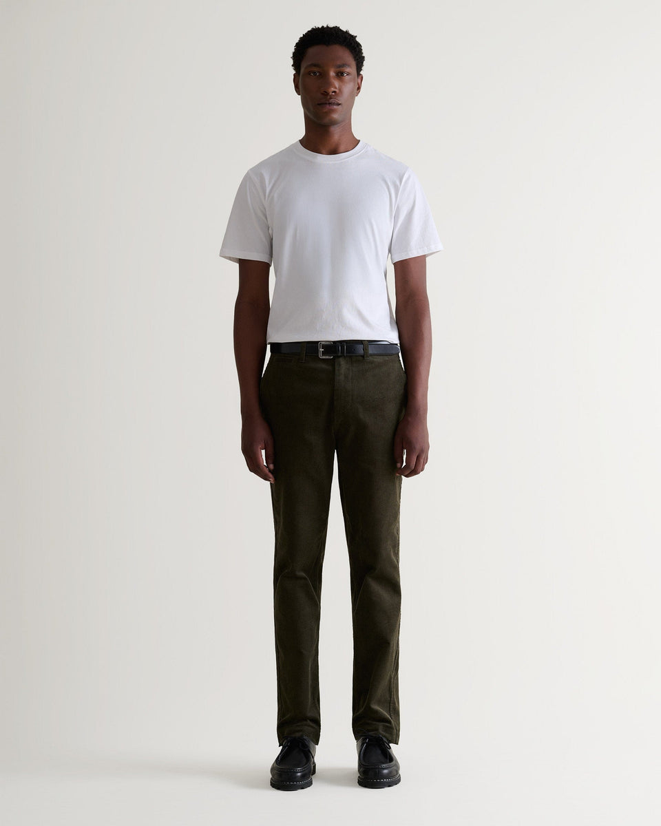 Full length front view of man wearing Rise & Fall's dark green corduroy trousers. Worn with white T-shirt and black shoes.