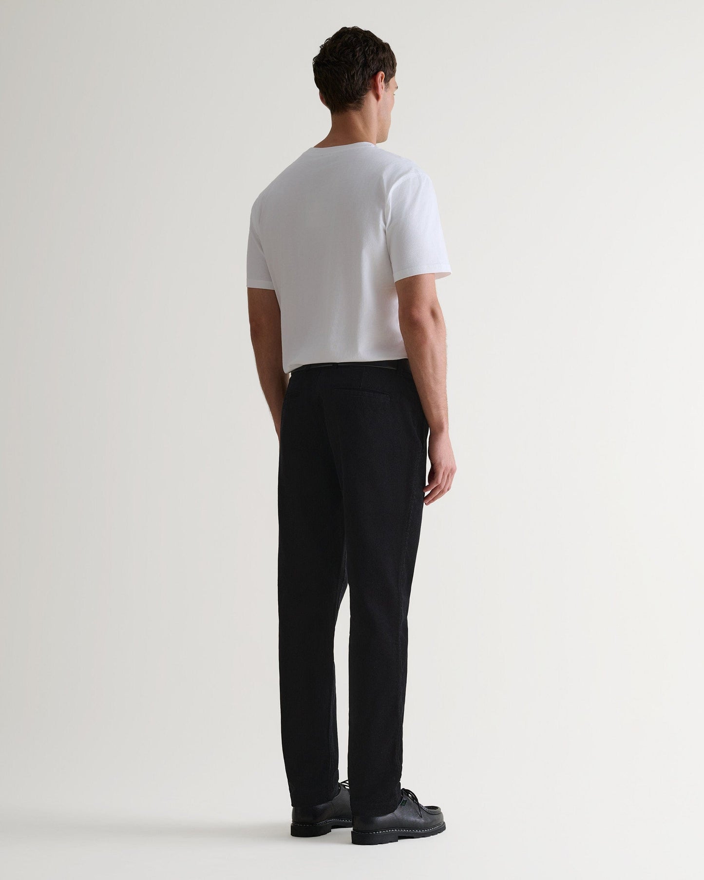 Back view of Rise & Fall's black corduroy trousers worn with black leather shoes and white T-shirt.