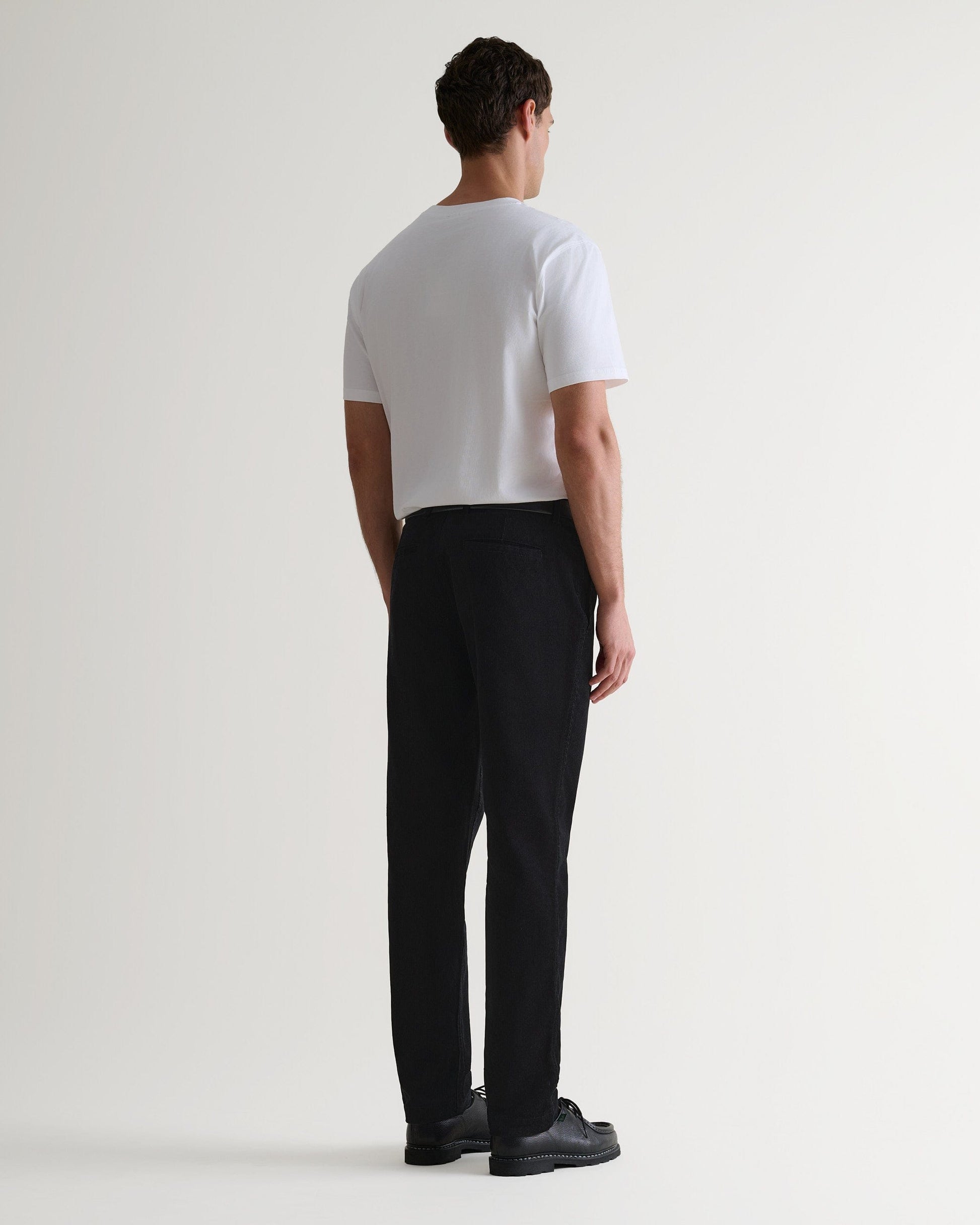 Back view of Rise & Fall's black corduroy trousers worn with black leather shoes and white T-shirt.