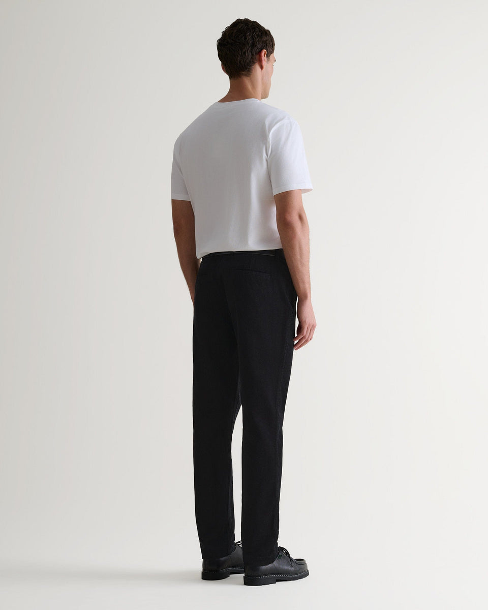 Back view of Rise & Fall's black corduroy trousers worn with black leather shoes and white T-shirt.