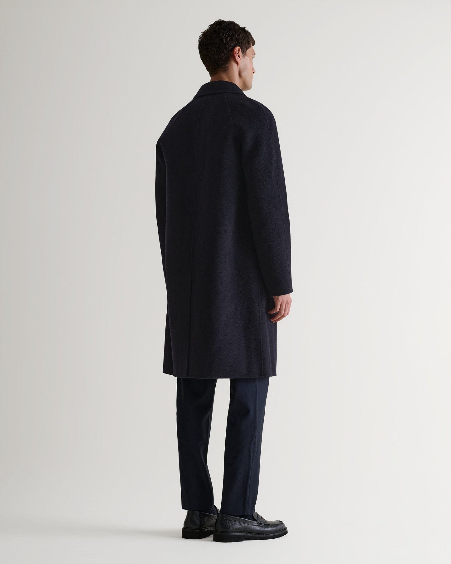 Full length back view of Rise & Fall's navy cashmere merino wool double-faced coat. Worn with black trousers and shoes.