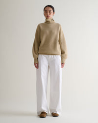 Front view of woman wearing beige cashmere merino Ottoman ribbed roll neck jumper with white jeans and tan suede shoes.