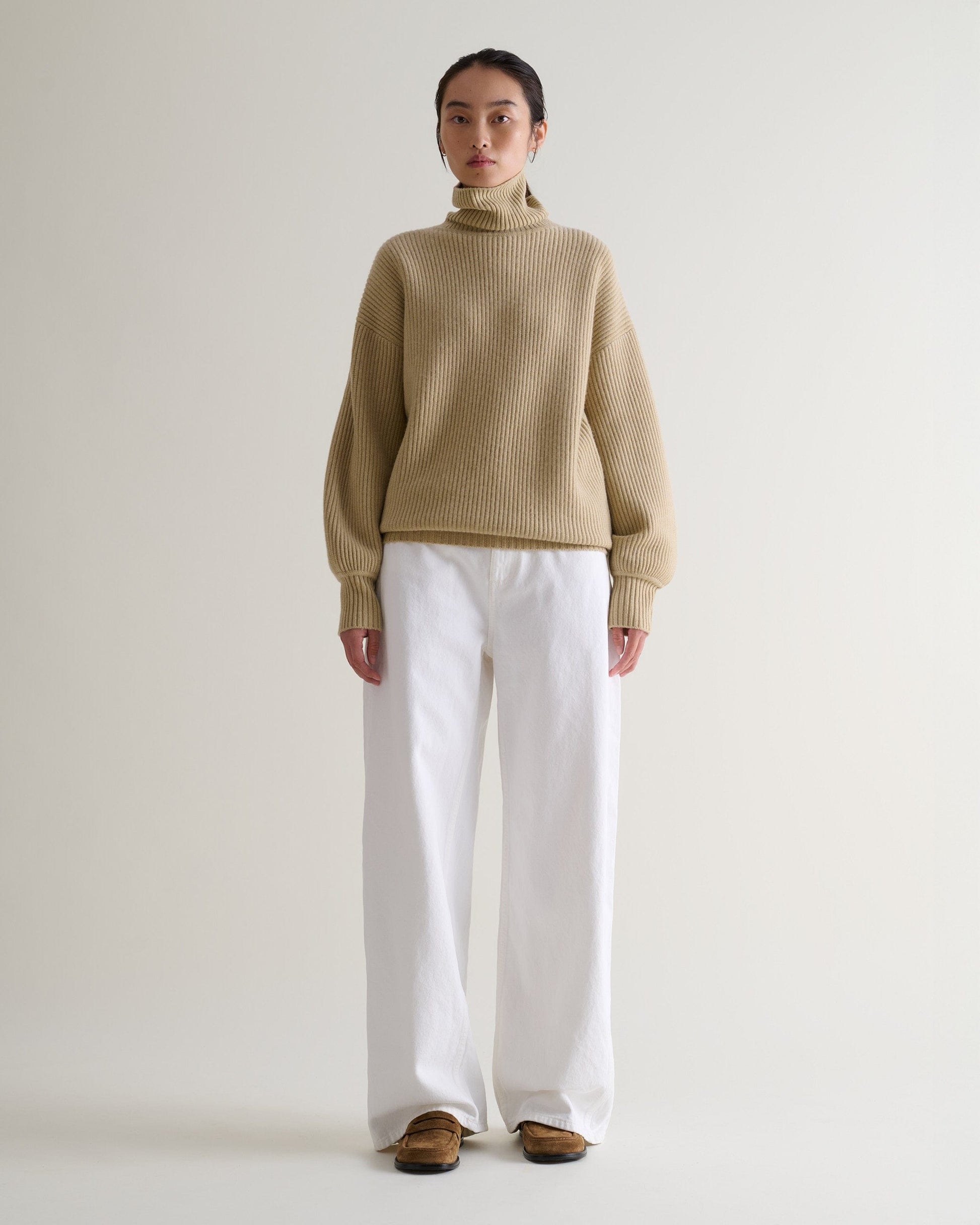 Front view of woman wearing beige cashmere merino Ottoman ribbed roll neck jumper with white jeans and tan suede shoes.