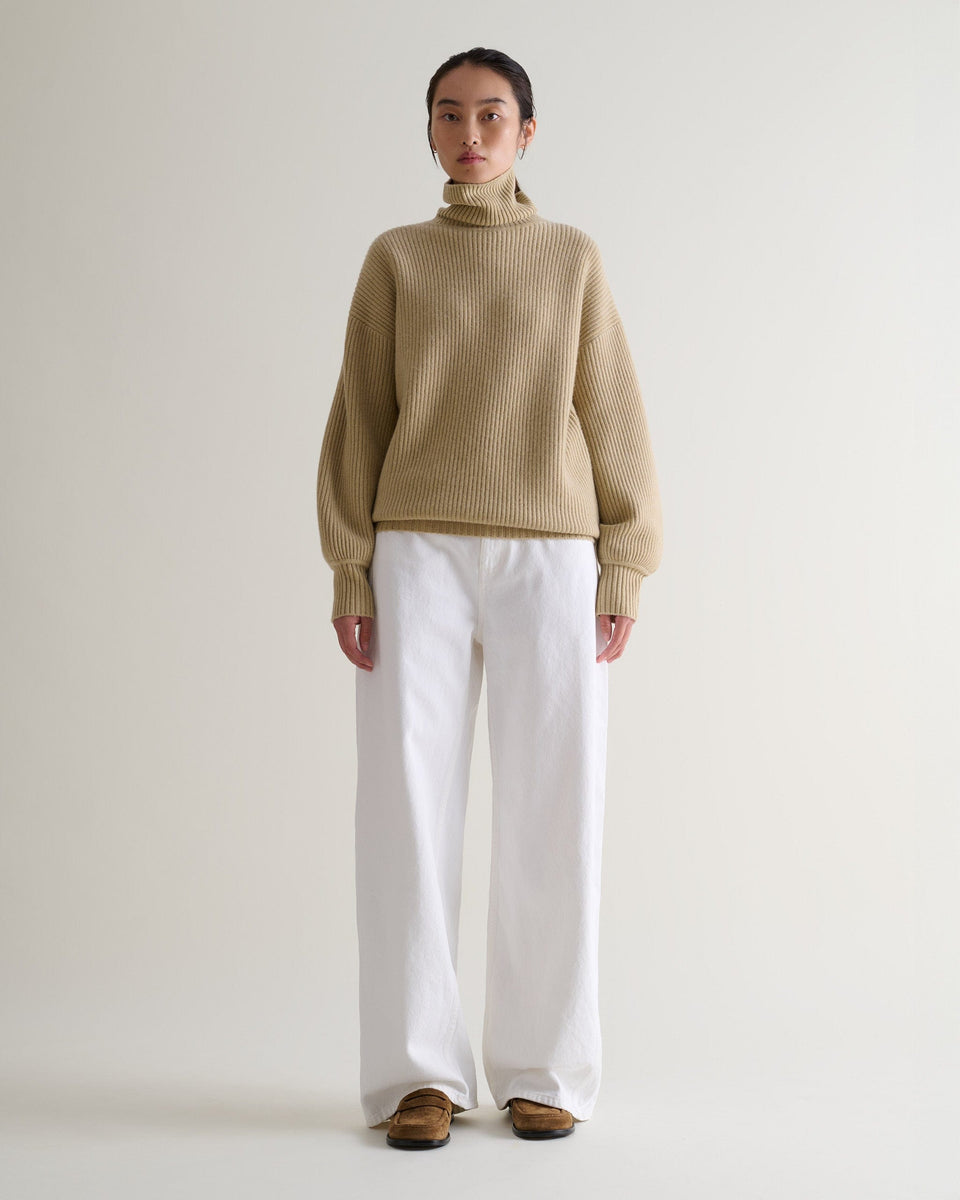 Front view of woman wearing beige cashmere merino Ottoman ribbed roll neck jumper with white jeans and tan suede shoes.