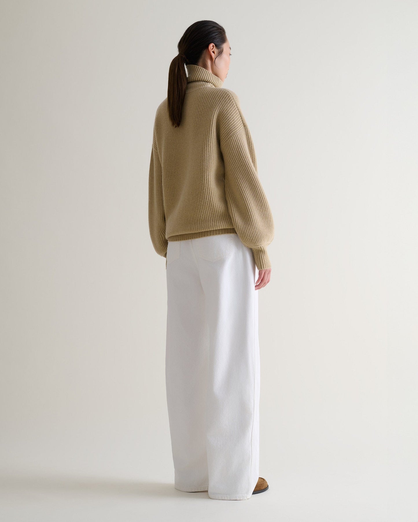 Back view of a woman wearing a beige Ottoman ribbed cashmere merino wool roll neck jumper with white jeans.