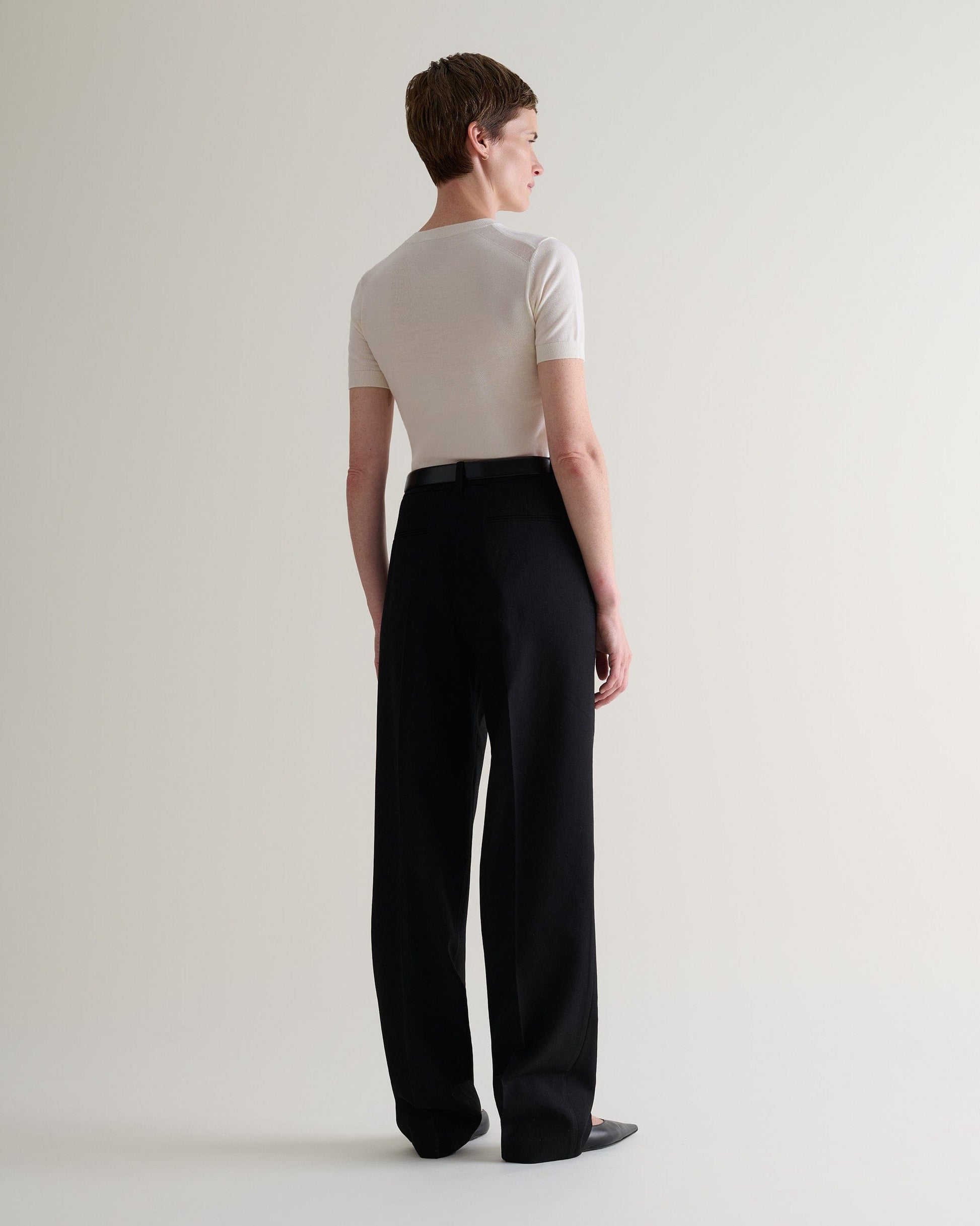 Full length back view of woman wearing cream superfine merino wool knitted t-shirt. Worn with black trousers and shoes.