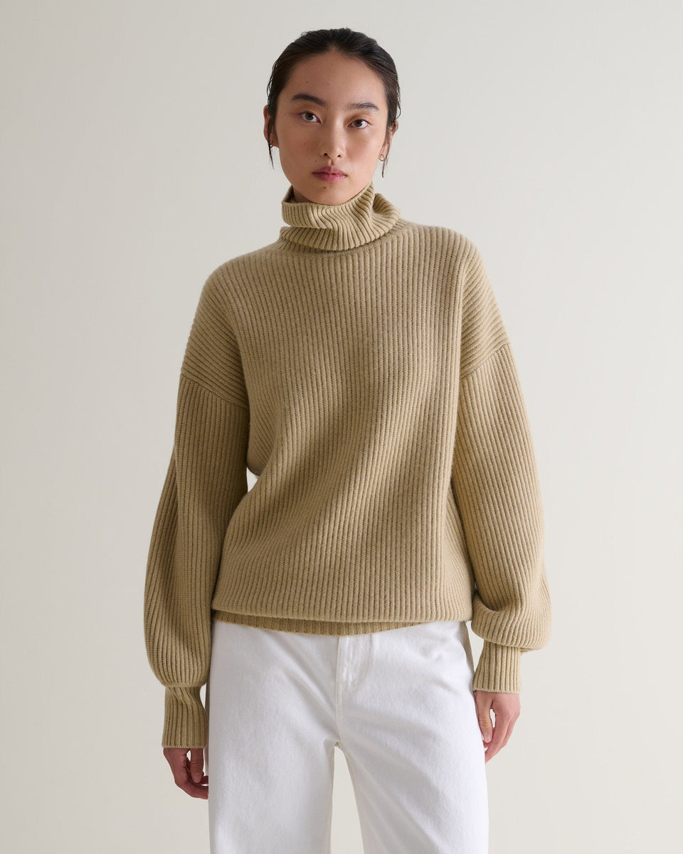 Close up front view of woman wearing twine beige cashmere merino wool Ottoman ribbed roll neck jumper with white trousers.