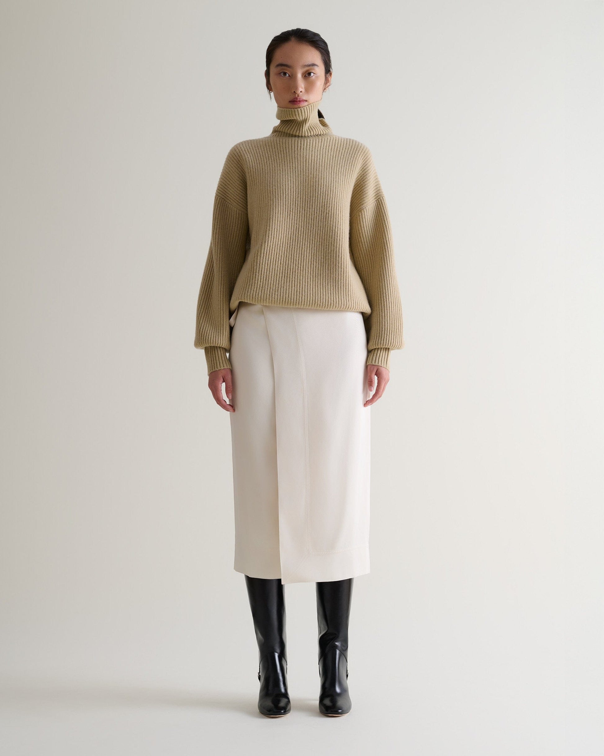 Woman wears a cream Naia™ wrap skirt with a beige cashmere roll neck jumper and black boots.