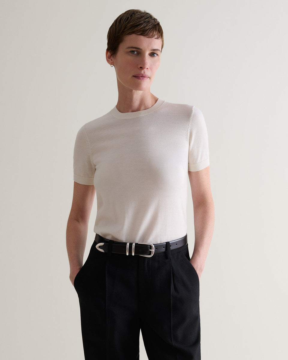Front view of woman wearing Rise & Fall's cream superfine merino wool knitted t-shirt. Worn with black trousers and belt.