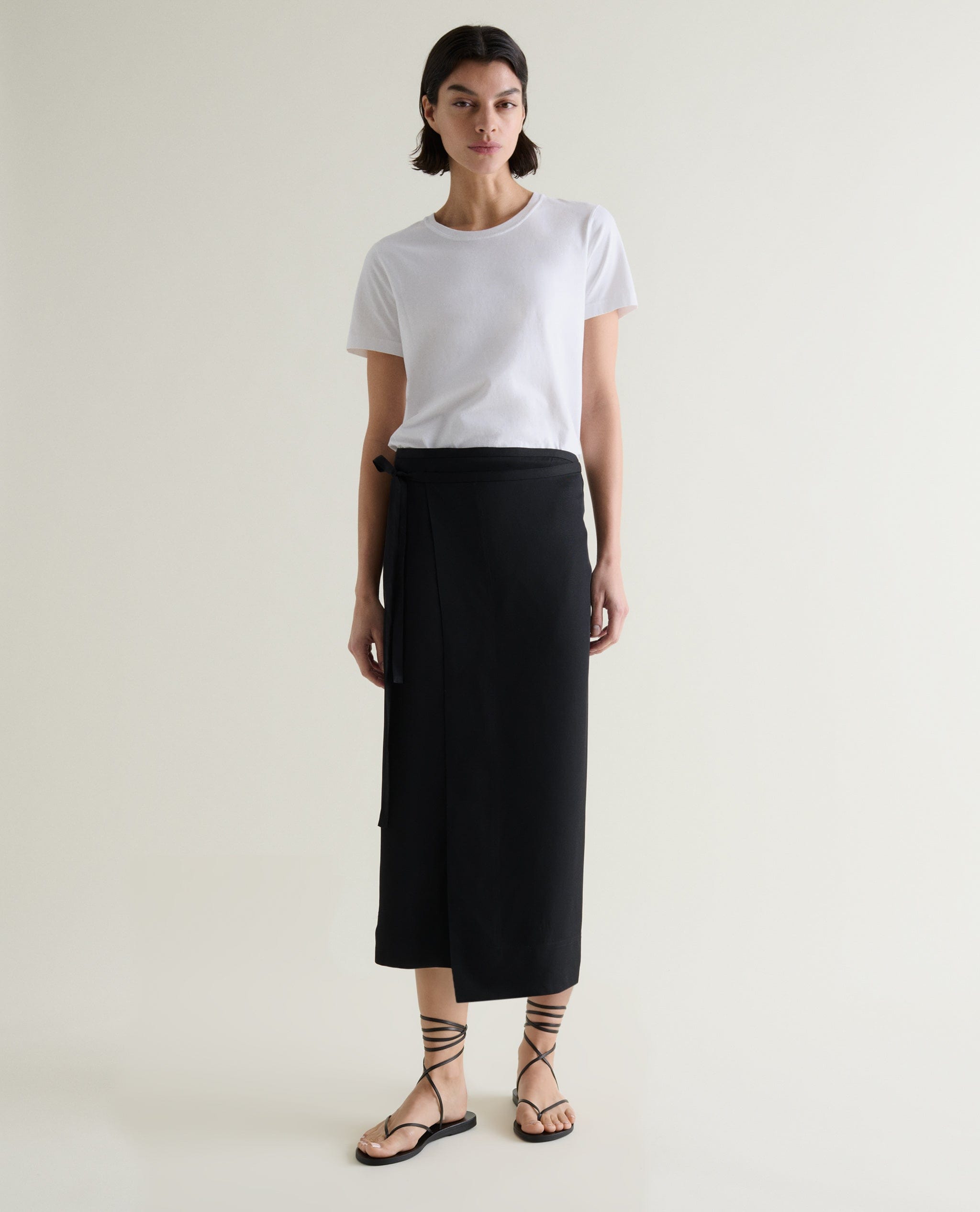 Woman wears Rise & Fall's black Naia™ skirt with a white classic T-shirt and black sandals. 