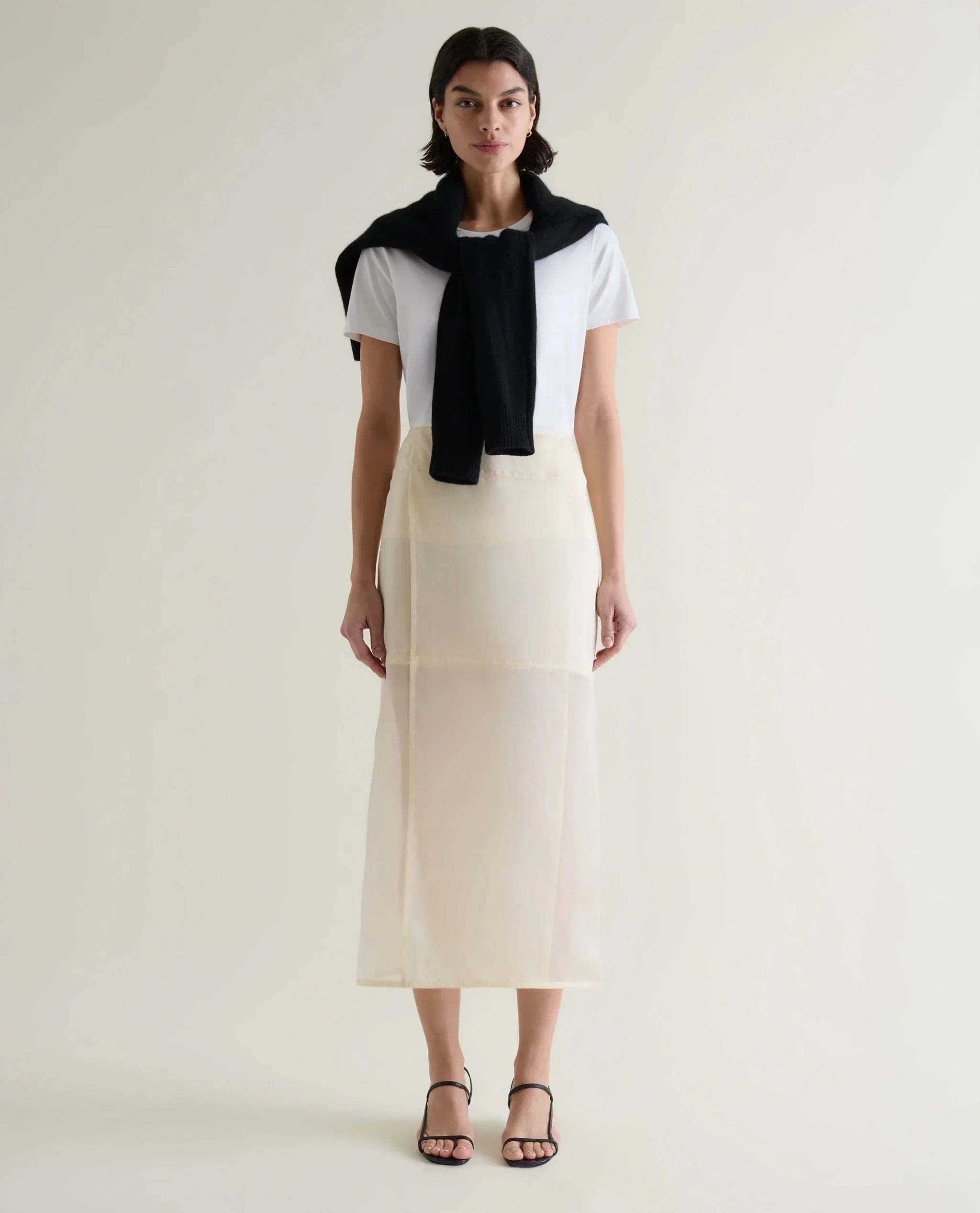 Woman wears silk organza panelled skirt in shell with a white T-shirt and black jumper around the shoulders. 